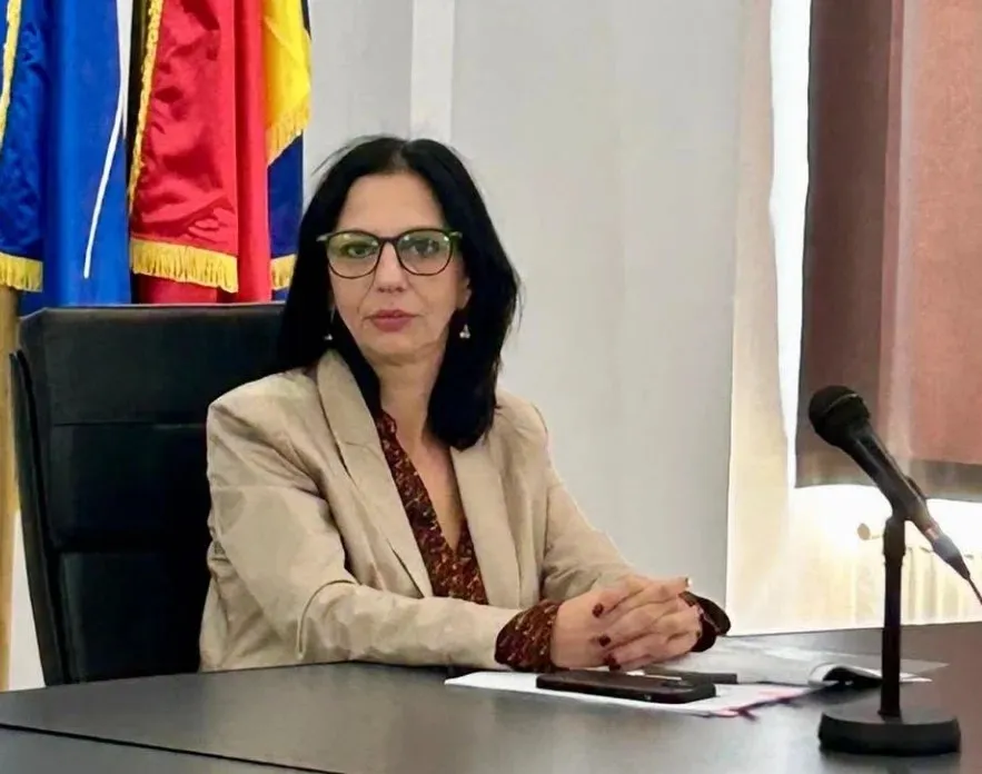 O femeie cu ochelari și păr negru stă la o masă lângă un microfon, purtând o jachetă bej peste o bluză închisă la culoare.