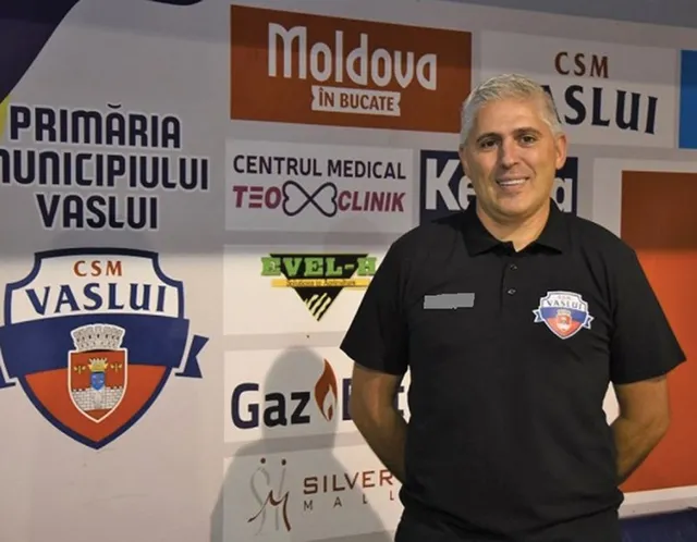 Un bărbat cu părul gri și tricou negru stă în fața unui perete cu diverse logouri și inscripții colorate.