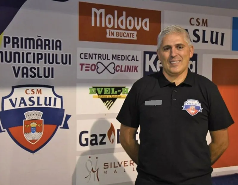 Un bărbat cu părul gri și tricou negru stă în fața unui perete cu diverse logouri și inscripții colorate.