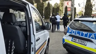 Două mașini de poliție sunt parcate pe un drum, iar trei persoane merg pe trotuar în direcția arborilor.