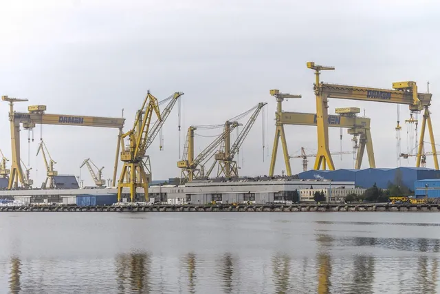 Mai multe macarale galben de tip portal și macarale de tip bra sunt amplasate pe malul unui port lângă câteva clădiri albe.