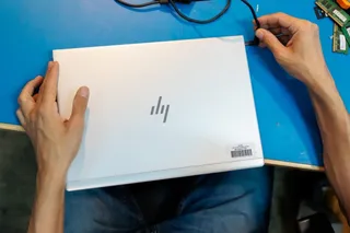Două mâini țin un laptop alb deschis pe o masă albastră lângă un cablu negru și componente electronice mici.