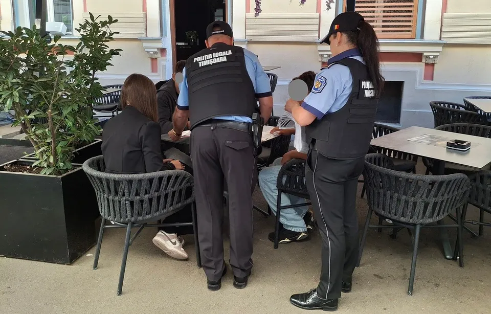 Unii polițiști în uniformă cu vestă tactică stau în picioare lângă o masă de cafea de la o terasă exterioară lângă un personaj așezat.