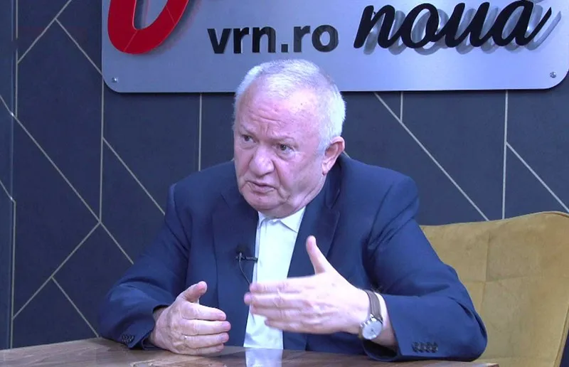 Un bărbat cu părul alb, îmbrăcat într-un sacou închis la culoare și cămașă albă, stă la o masă și gesticulează cu mâinile.