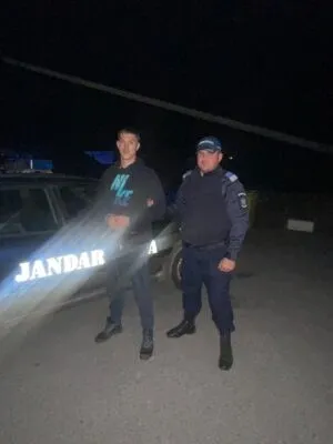 Un bărbat cu hanorac negru și un agent de poliție în uniformă albastră stau lângă un vehicul marcat cu textul JANDARM.