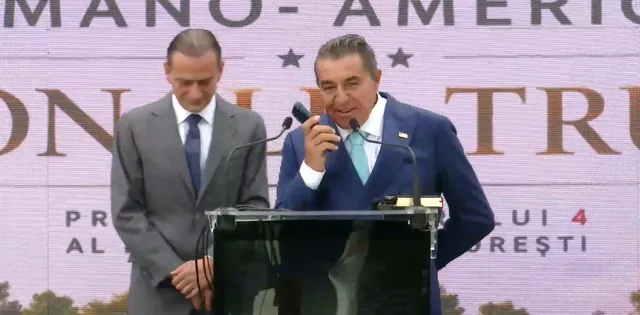 Un bărbat într-un costum albastru vorbește la un microfon de la un podium, în timp ce un alt bărbat într-un costum gri stă lângă el.