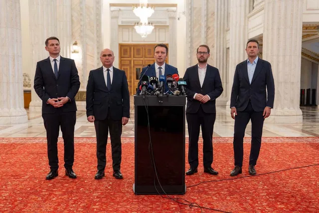 Cinci bărbați în costume stau în picioare pe un covor roșu în fața unui podium cu microfoane într-o încăpere cu coloane albe.