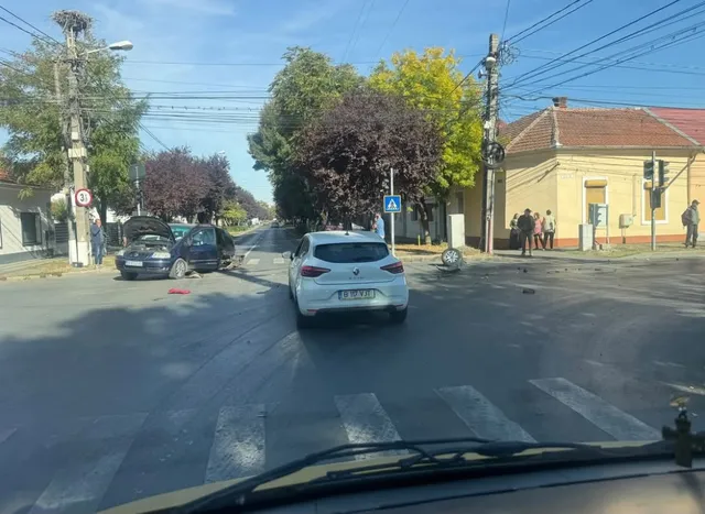 Un autoturism alb este oprit pe un drum asfaltat lângă un zebru, în timp ce două mașini de culoare închisă sunt poziționate în lateral.