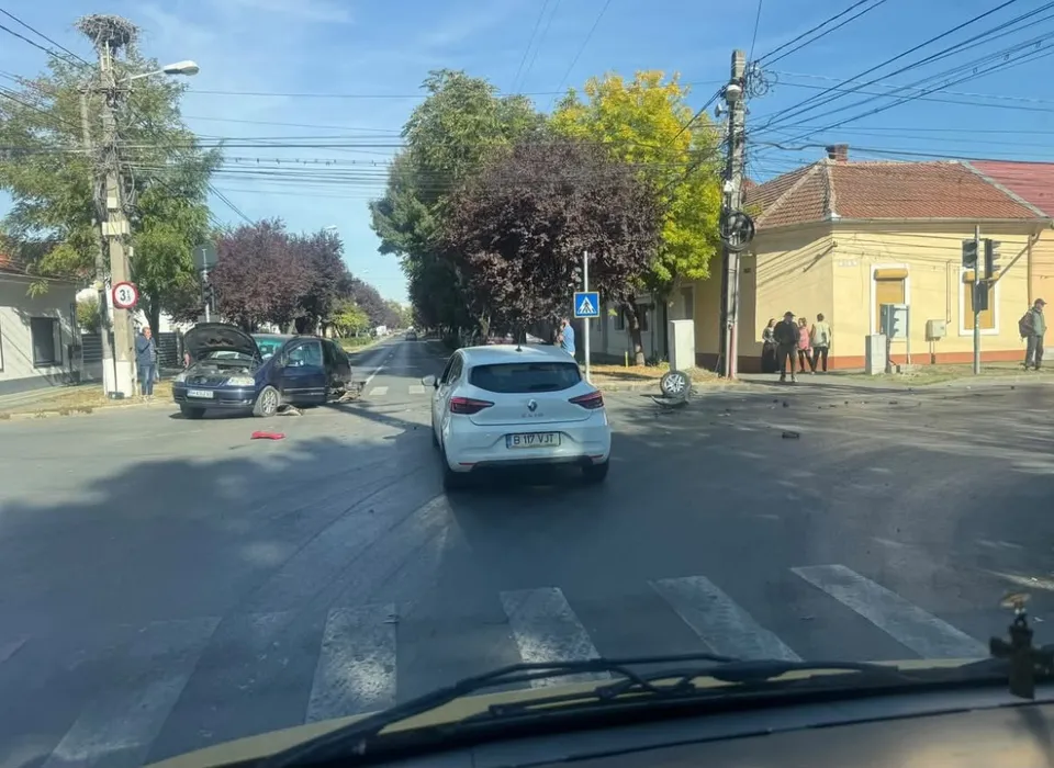 Un autoturism alb este oprit pe un drum asfaltat lângă un zebru, în timp ce două mașini de culoare închisă sunt poziționate în lateral.
