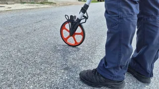 O persoană în pantaloni albaștri și încălțăminte neagră stă lângă un obiect cu o roată portocalie pe un drum asfaltat.