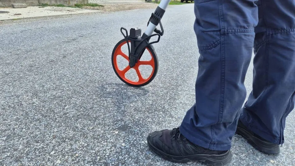 O persoană în pantaloni albaștri și încălțăminte neagră stă lângă un obiect cu o roată portocalie pe un drum asfaltat.