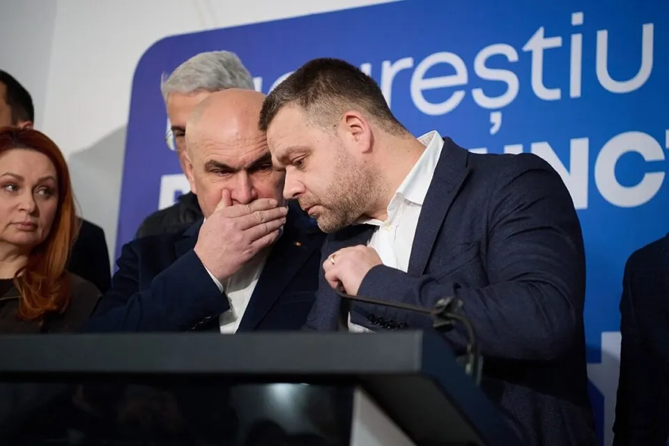 Trei bărbați și o femeie stau în spatele unui podium, în timp ce un bărbat în costum închis își acoperă gura cu mâna.