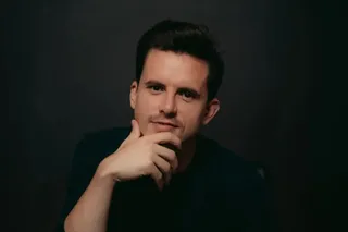 Un bărbat cu părul scurt și întunecat stă cu mâna sub bărbie în fața unui fundal negru.