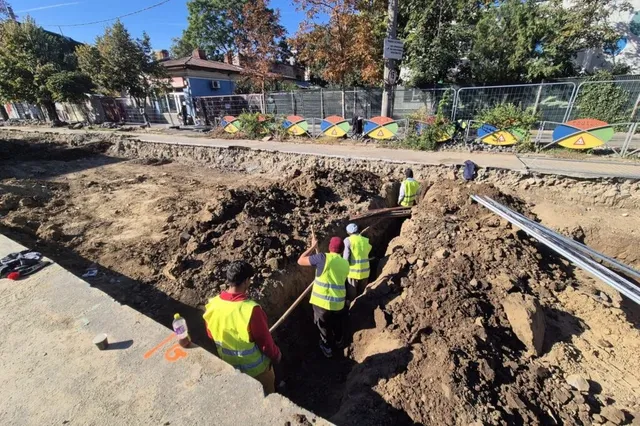 Câțiva muncitori cu veste reflectorizante de culoare galbenă lucrează într-o șanț adânc excavat în pământ lângă un trotuar.