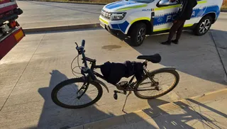 O bicicletă neagră este așezată pe un trotuar lângă o mașină de poliție albă și albastră parcată pe asfalt.