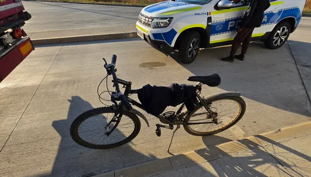 O bicicletă neagră este așezată pe un trotuar lângă o mașină de poliție albă și albastră parcată pe asfalt.