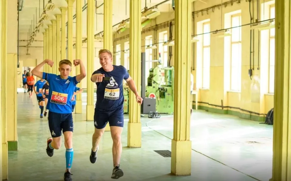 Doi bărbați în echipament sportiv de culoare albastră aleargă printr-un spațiu industrial cu coloane galbene și ferestre înalte.