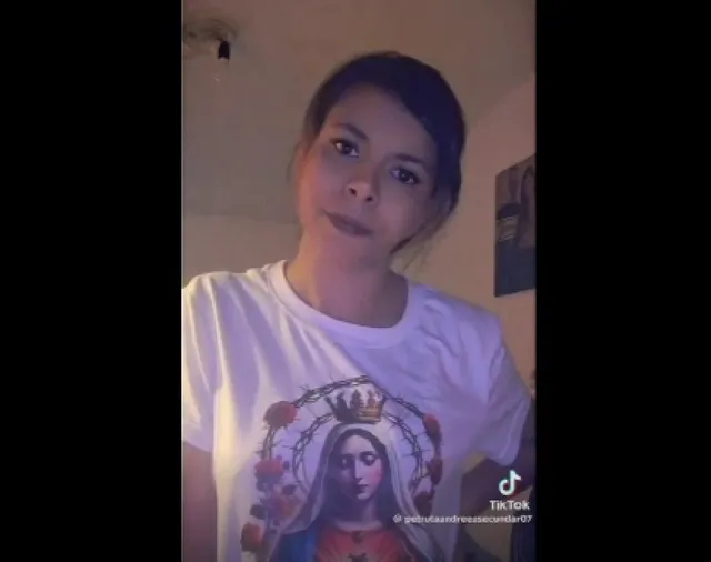 O femeie cu părul închis la culoare poartă un tricou alb cu o reprezentare grafică a unei figuri religioase sub o coroană.