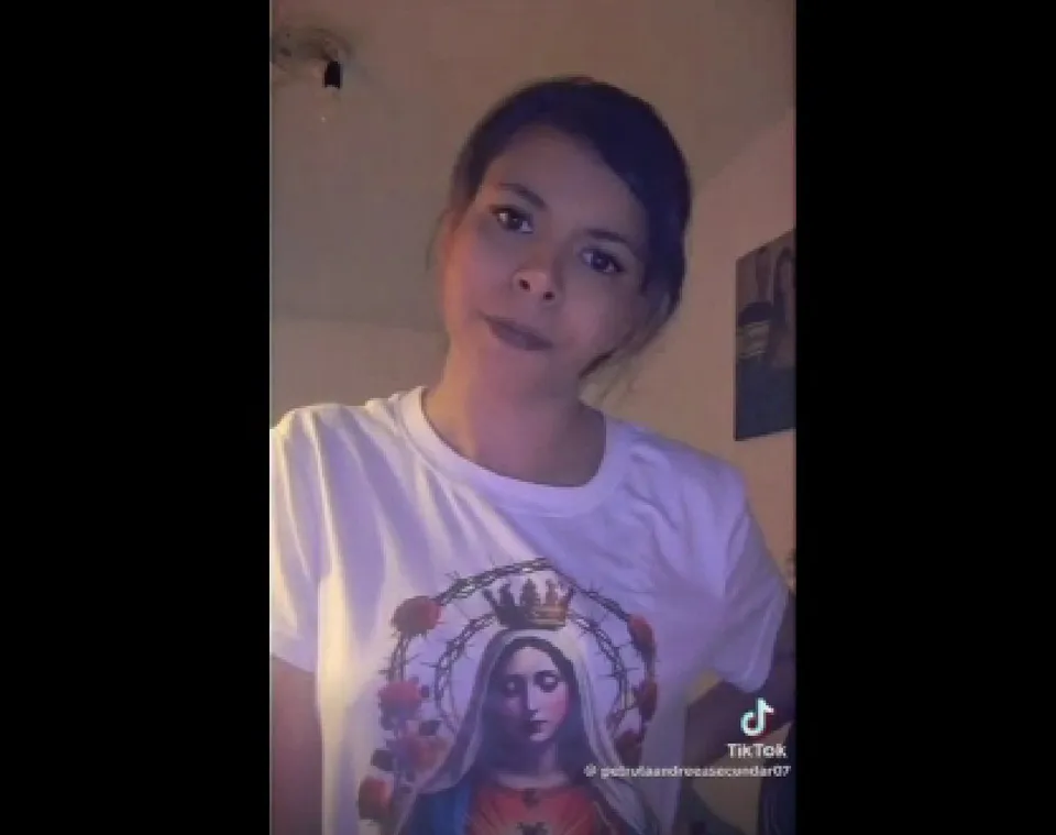 O femeie cu părul închis la culoare poartă un tricou alb cu o reprezentare grafică a unei figuri religioase sub o coroană.
