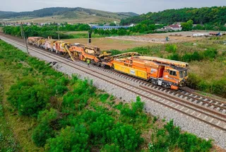 Un tren de lucru format din mai multe vagoane portocalii și galbene circulă pe o cale ferată într-un peisaj verde cu dealuri în fundal.