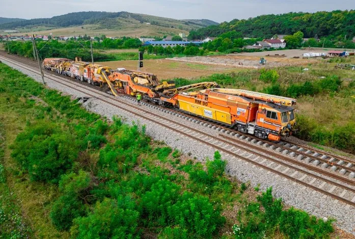 Un tren de lucru format din mai multe vagoane portocalii și galbene circulă pe o cale ferată într-un peisaj verde cu dealuri în fundal.