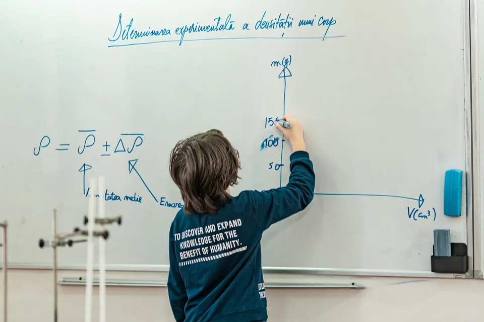 Un copil cu părul șaten scrie formule matematice pe o tablă albă mare într-o încăpere luminată.