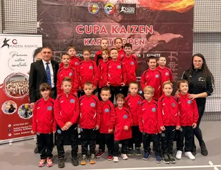 Un grup de copii în echipamente sportive roșii și negre posează pentru o fotografie alături de doi adulți în fața unui panou publicitar.