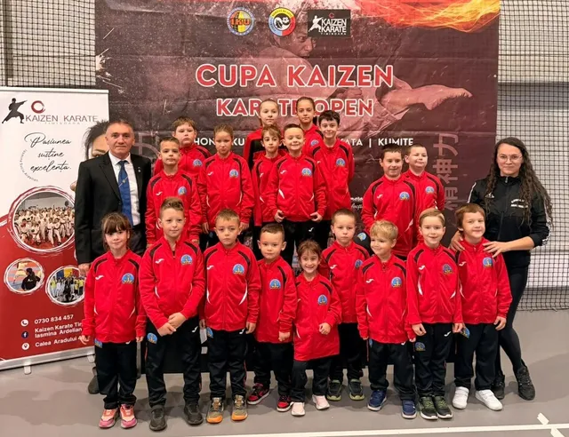 Un grup de copii în echipamente sportive roșii și negre posează pentru o fotografie alături de doi adulți în fața unui panou publicitar.