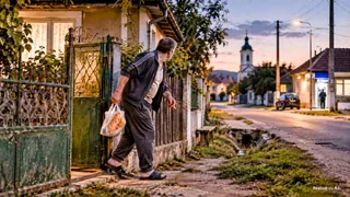 Un bărbat cu o pungă de pâine în mână pășește pe un trotuar lângă o poartă metalică verde și un gard de lemn.