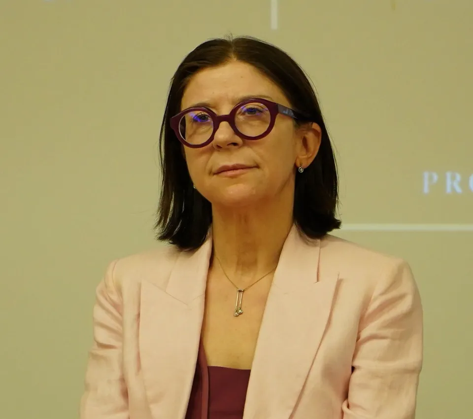 O femeie cu ochelari cu rame roșii și un blazer roz poartă un colier subțire cu un pandantiv argintiu.