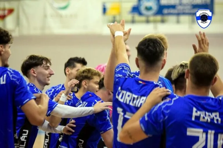 Un grup de sportivi îmbrăcați în echipamente albastre de handbal stau împreună într-un grup, cu brațele ridicate.