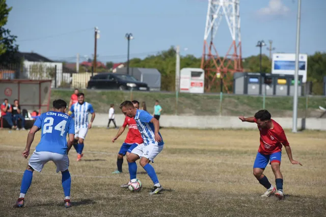 Câțiva jucători de fotbal în echipamente albastre și roșii joacă pe un teren de iarbă uscată sub un cer senin.