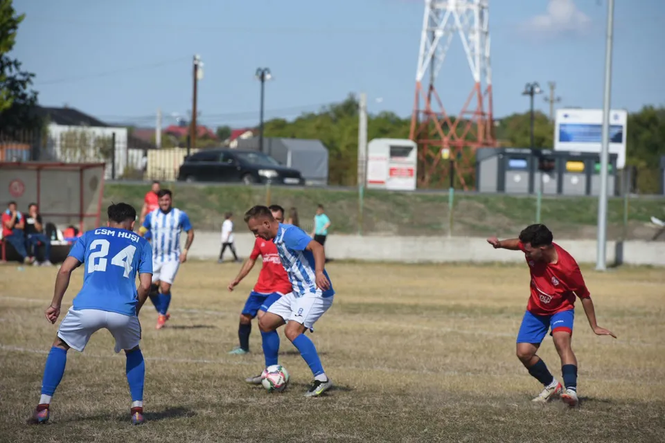 Câțiva jucători de fotbal în echipamente albastre și roșii joacă pe un teren de iarbă uscată sub un cer senin.