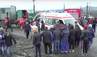 Un grup de oameni stă pe un teren de noroi lângă o ambulanță albă și un camion de pompieri într-un spațiu deschis.