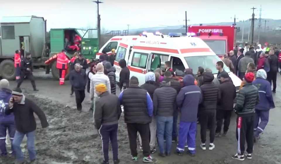 Un grup de oameni stă pe un teren de noroi lângă o ambulanță albă și un camion de pompieri într-un spațiu deschis.