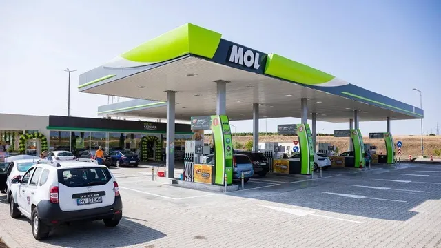 O benzinărie cu un acoperiș mare gri și verde este situată lângă un drum, având mai multe mașini parcate sub copertă.