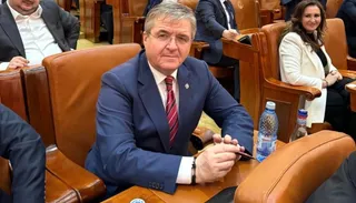Un bărbat cu ochelari și costum albastru stă așezat într-un fotoliu de piele maro, ținând un pix în mână.