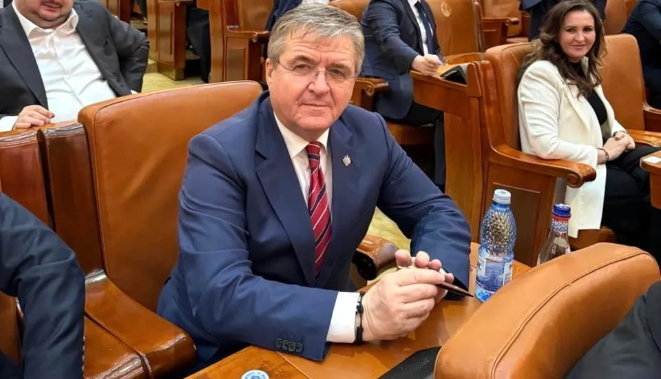 Un bărbat cu ochelari și costum albastru stă așezat într-un fotoliu de piele maro, ținând un pix în mână.