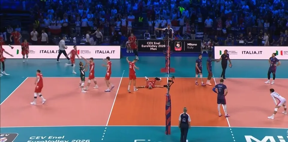 Jucători de volei în echipamente roșii și albastre participă la un meci pe un teren de culoare portocalie și verde în fața unei tribune.