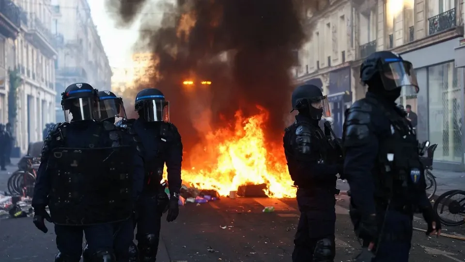 Patru polițiști în echipament de protecție și cască stau pe un drum urban lângă un foc de tabără cu fum negru dens.