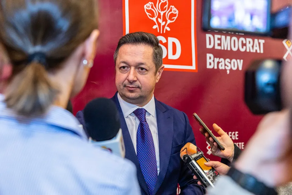 Un bărbat într-un costum albastru și cravată violet este intervievat de mai mulți oameni care țin microfoane și un telefon mobil în față.