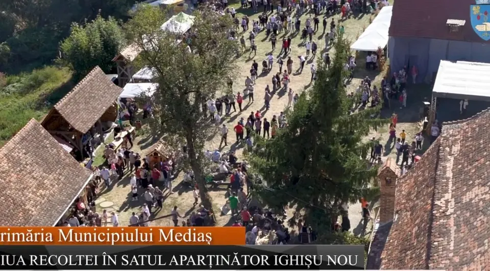 O mulțime de oameni este adunată într-o zonă deschisă între clădiri și copaci, sub lumina soarelui.