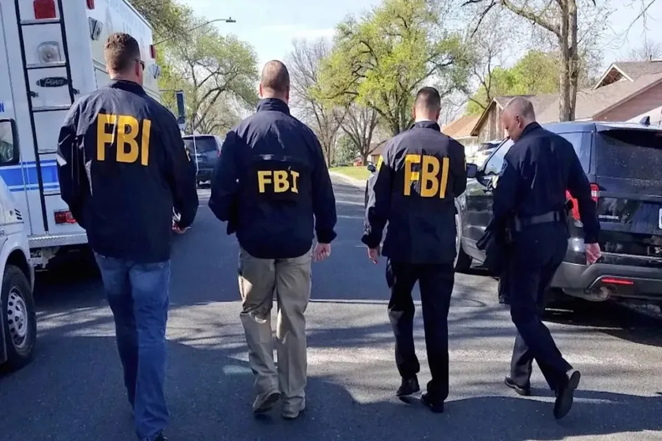 Patru bărbați merg pe un drum lângă mașini, fiind doi dintre ei purtând jachete negre cu inscripția FBI scrisă în litere galbene pe spate.