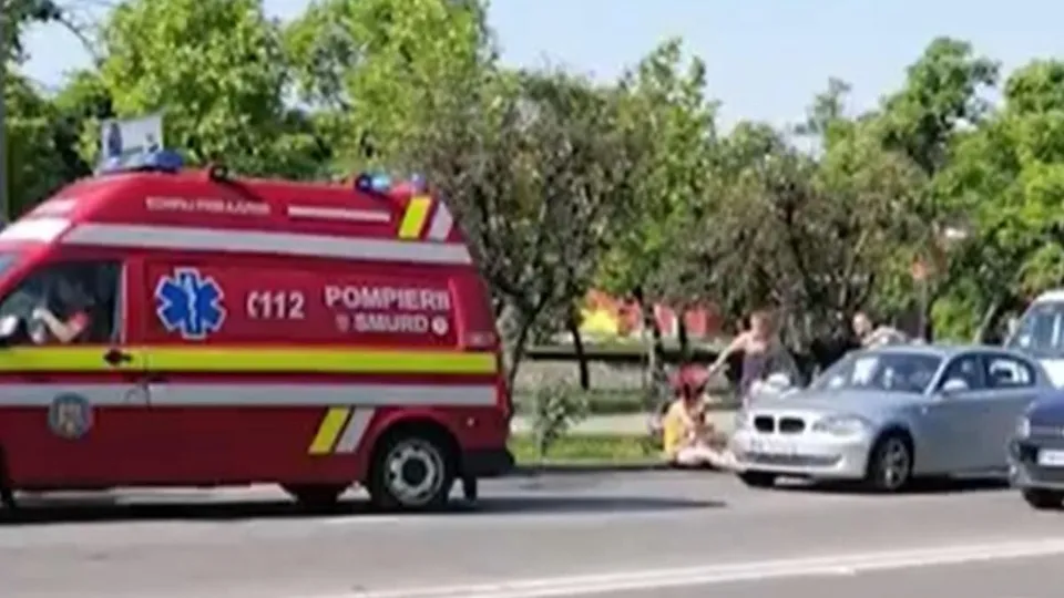 O ambulanță roșie cu inscripții este parcată pe marginea drumului lângă un autoturism argintiu și persoane aflate pe trotuar.