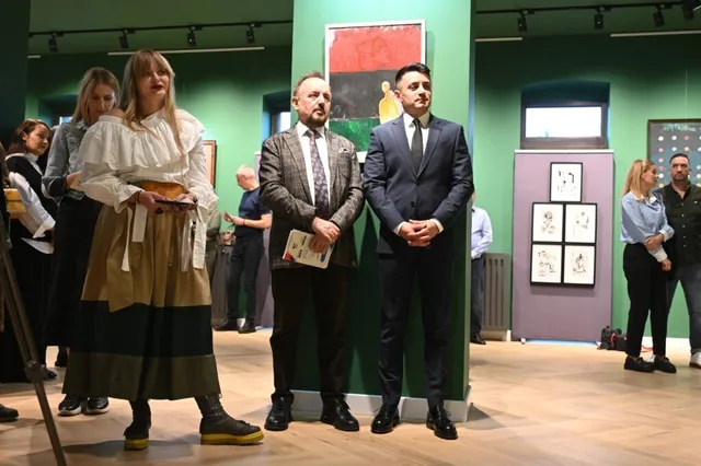 Cinci persoane stau în picioare într-o încăpere cu pereți verzi, în timp ce pe fundal sunt expuse diverse tablouri pe panouri albe.