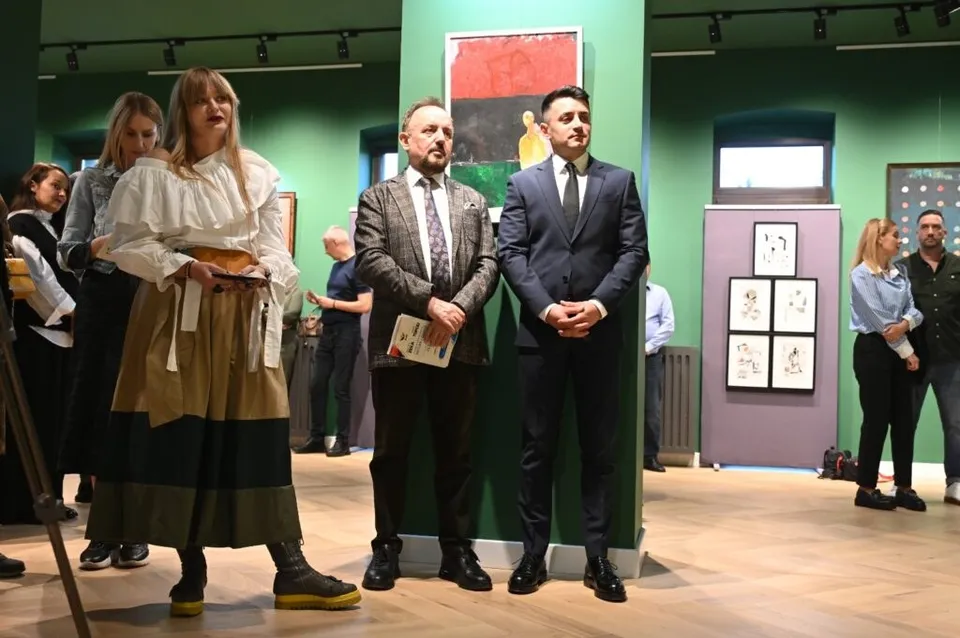 Cinci persoane stau în picioare într-o încăpere cu pereți verzi, în timp ce pe fundal sunt expuse diverse tablouri pe panouri albe.