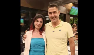 Un bărbat într-un tricou polo galben stă lângă o femeie cu părul roșcat purtând un sacou deschis și o rochie albastră.