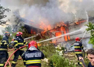 Patru pompieri în uniforme de lucru și casci colorați direcționează jeturi de apă către o clădire în flăcări din care se ridică fum dens.
