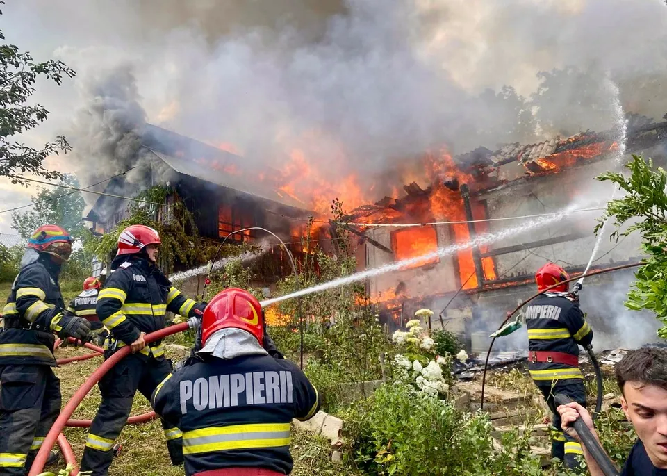 Patru pompieri în uniforme de lucru și casci colorați direcționează jeturi de apă către o clădire în flăcări din care se ridică fum dens.