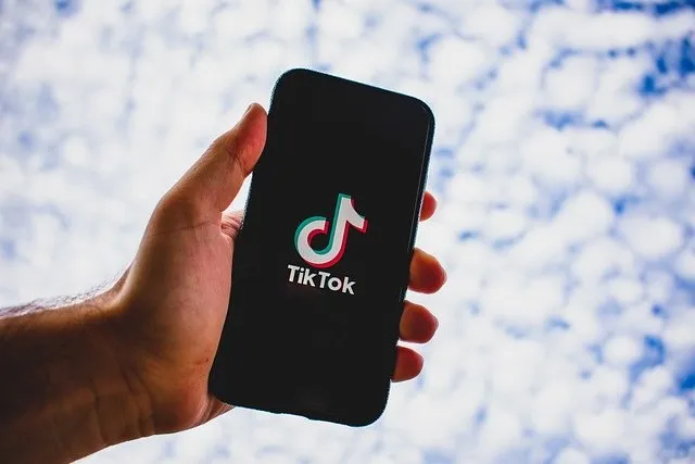 O mână ține un smartphone negru care afișează logo-ul TikTok pe un fundal de cer cu nori albi.